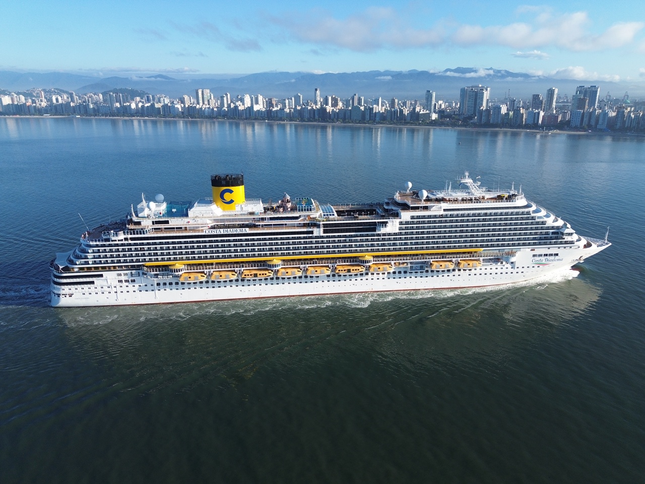 Vista aérea lateral de um grande navio da Costa Cruzeiros navegando na baía, com a orla e os altos edifícios da cidade de Santos ao fundo