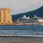 Navio de cruzeiro no horizonte navegando no mar visto a partir da faixa de areia, com edifícios residenciais altos à esquerda da orla de Santos