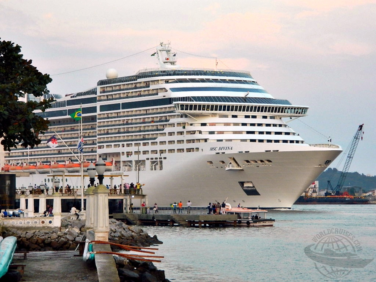 Navio da MSC Cruzeiros (classe Fantasia) navegando pelo canal do porto, visto a partir de um píer de pedras com pessoas observando próximos à mureta