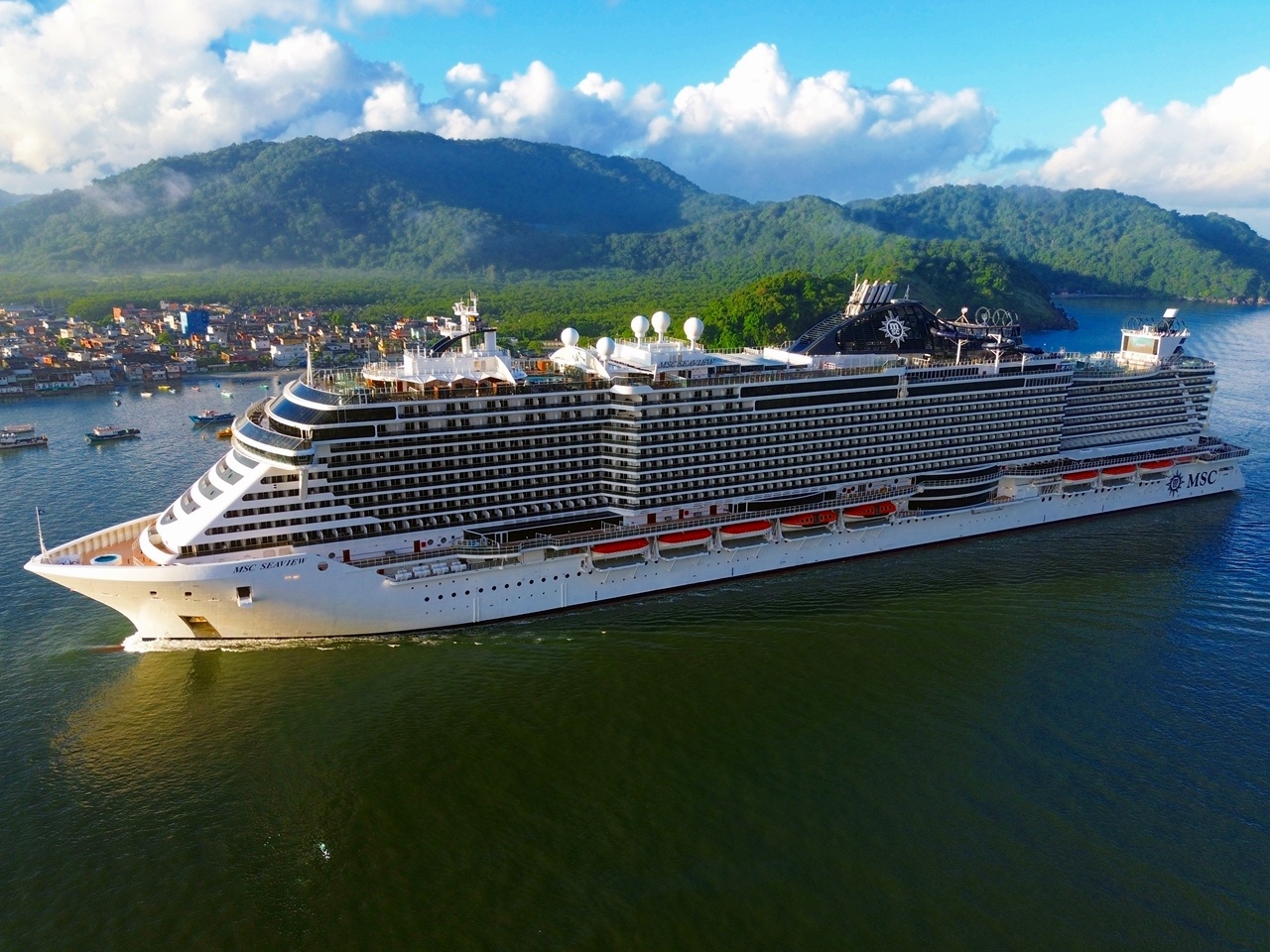 Vista aérea de um navio da MSC Cruzeiros (classe Seaside) navegando em águas costeiras, com morros cobertos por vegetação verde ao fundo