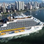 Vista aérea lateral de um grande navio da Costa Cruzeiros navegando na baía, com a orla e os altos edifícios da cidade de Santos ao fundo