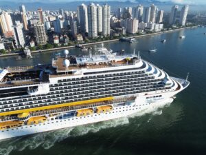 Vista aérea lateral de um grande navio da Costa Cruzeiros navegando na baía, com a orla e os altos edifícios da cidade de Santos ao fundo