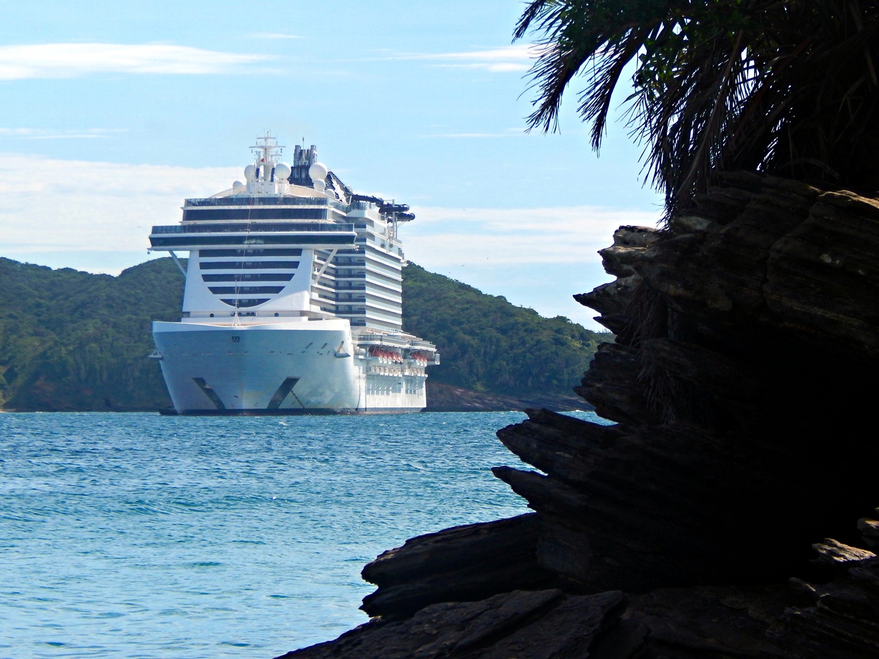 Navio de cruzeiro da classe Seaside (MSC) visto de frente, emoldurado por rochas e vegetação costeira à direita da imagem em Búzios