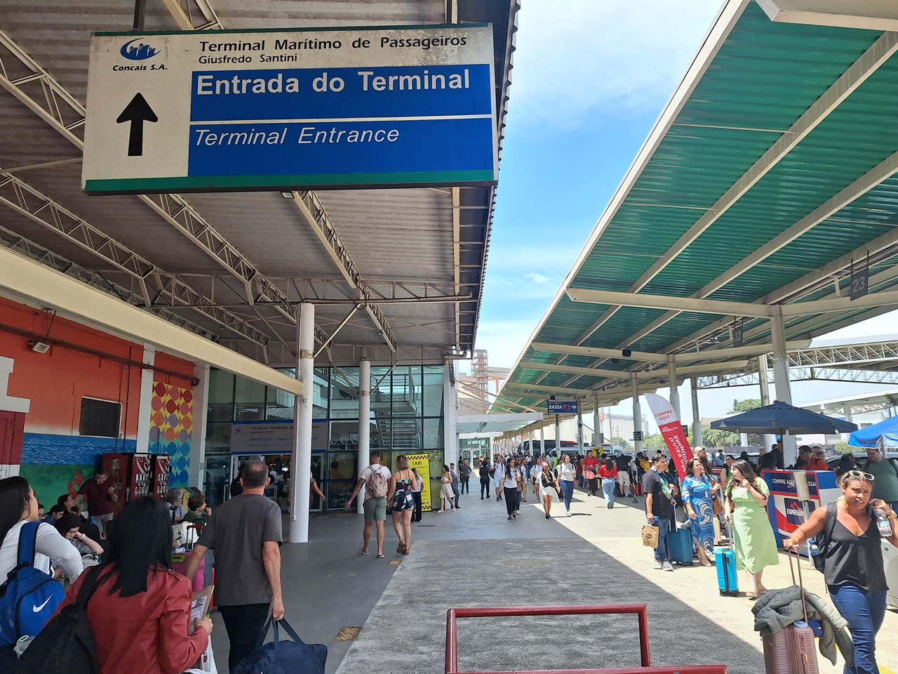 Movimento de passageiros com malas caminhando sob a estrutura coberta na área externa do Terminal Concais, com uma placa azul suspensa indicando a "Entrada do Terminal"