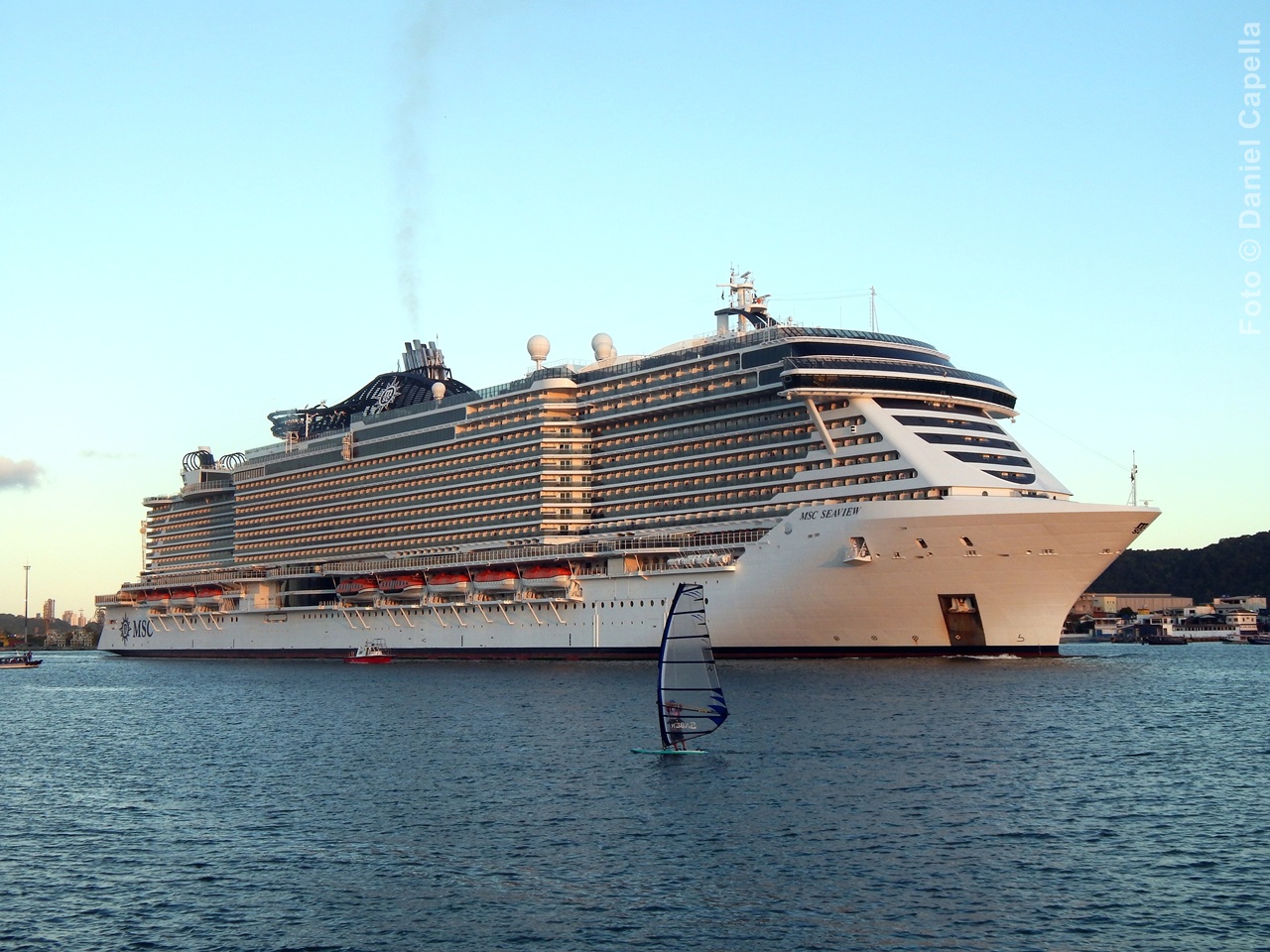 Navio MSC Seaview visto de perfil navegando, com destaque para sua arquitetura traseira aberta e um praticante de windsurf no mar em primeiro plano