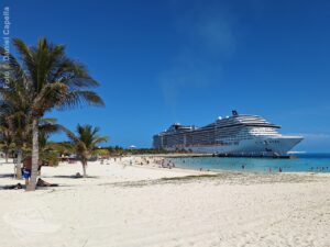 Areia branca e coqueiros em primeiro plano, com o MSC Divina ao fundo durante escala em Ocean Cay