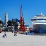 Fotografia diurna da Praça Mauá no Rio de Janeiro. À direita, a proa de um grande navio da Costa Cruzeiros atracado no porto. Ao centro e à esquerda, destacam-se dois antigos guindastes portuários pintados de vermelho, o prédio histórico de torre branca do terminal e pedestres caminhando pelo calçadão sob céu azul