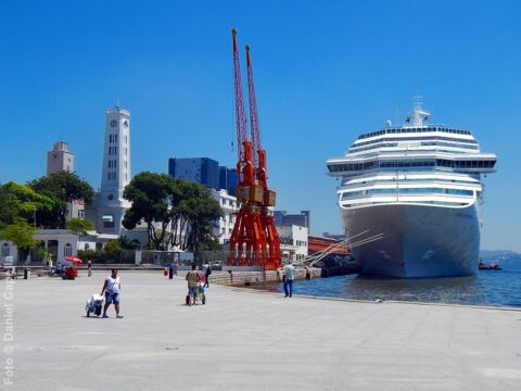 Fotografia diurna da Praça Mauá no Rio de Janeiro. À direita, a proa de um grande navio da Costa Cruzeiros atracado no porto. Ao centro e à esquerda, destacam-se dois antigos guindastes portuários pintados de vermelho, o prédio histórico de torre branca do terminal e pedestres caminhando pelo calçadão sob céu azul