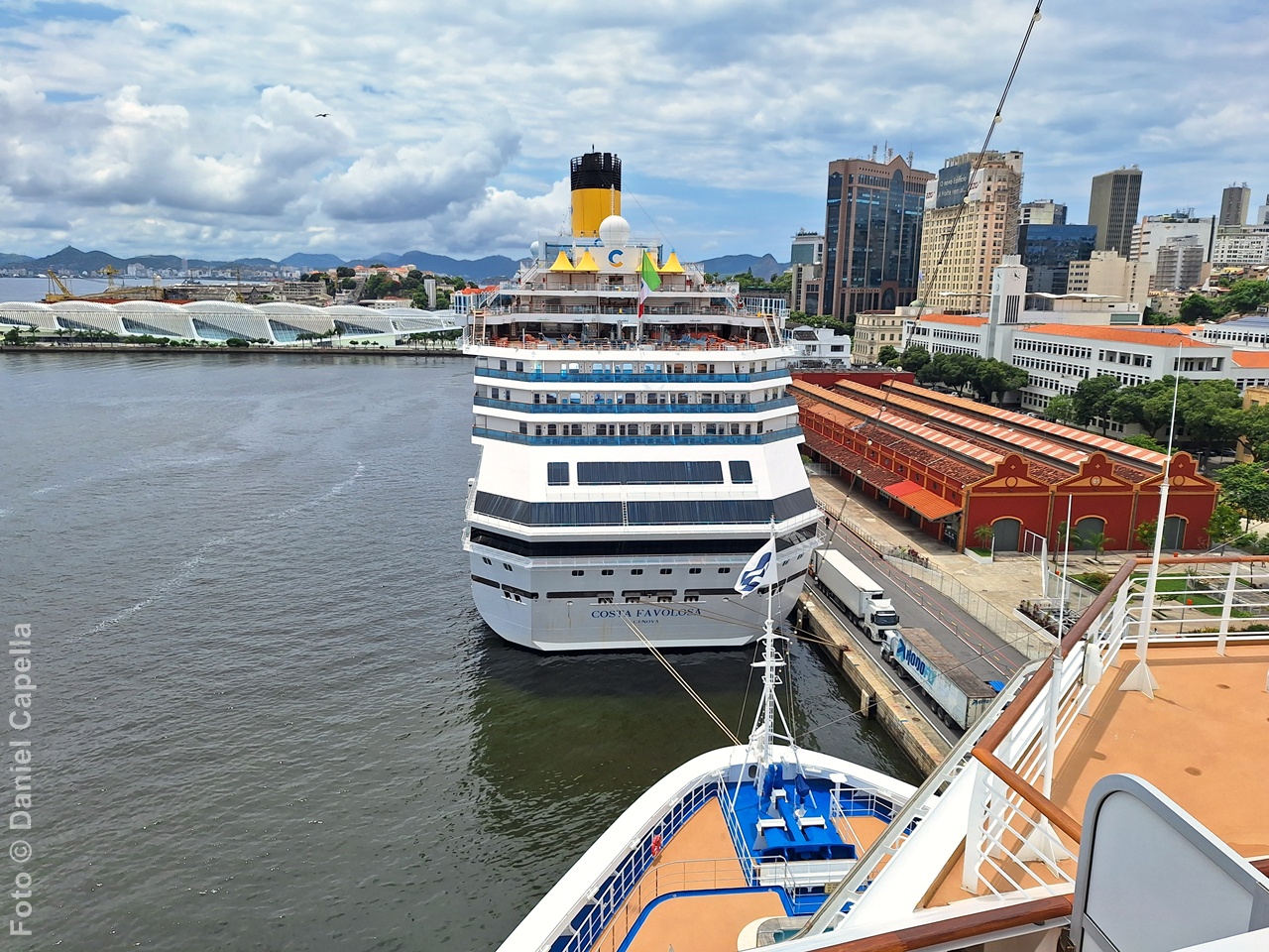 Fotografia de um navio da Costa Cruzeiros atracado no Pier Mauá, no Rio de Janeiro. À direita, aparecem os armazéns vermelhos do porto e, ao fundo à esquerda, o Museu do Amanhã e os prédios do centro da cidade sob um céu azul com nuvens